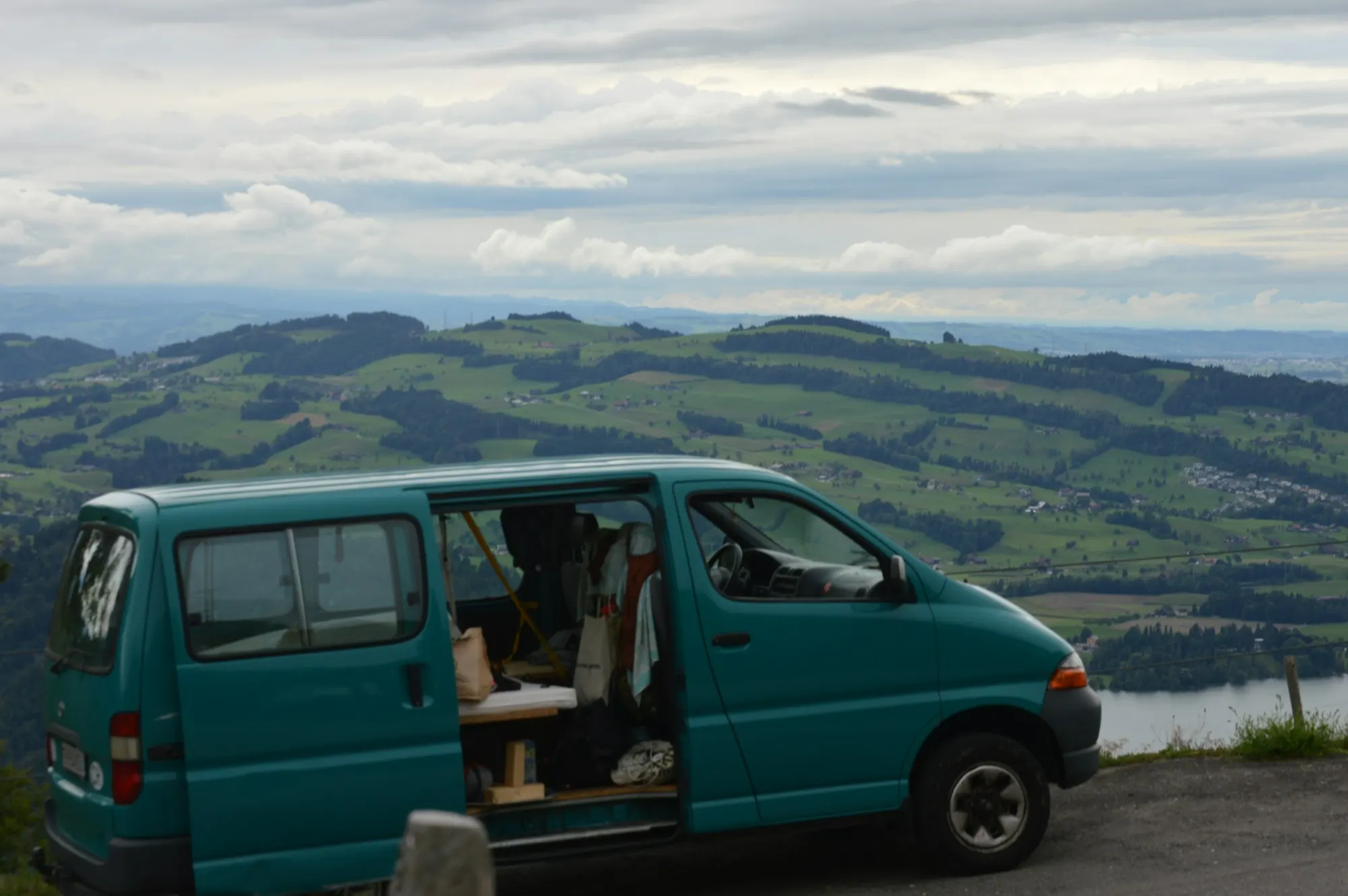 Teal campervan parked on a ridge above a mountain lake, side door open